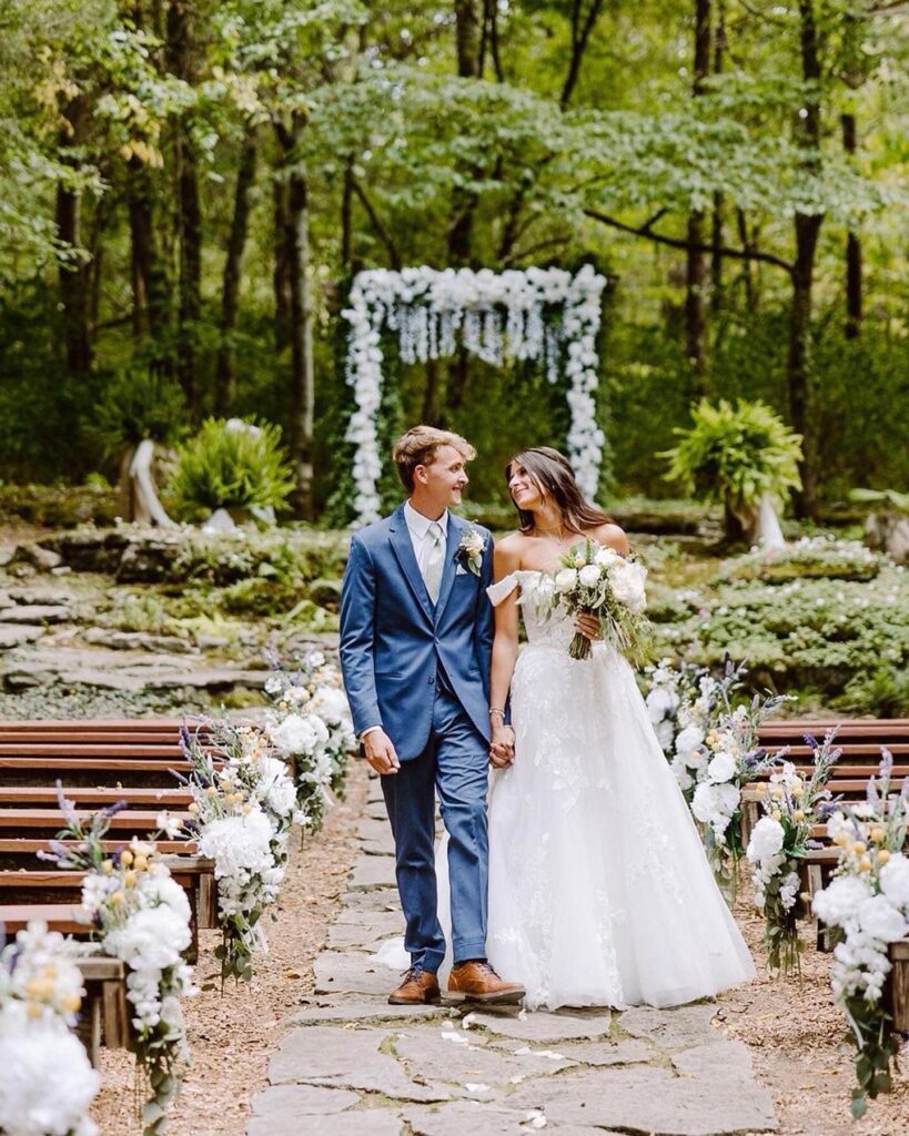 About 1 bride and groom walking down aisle in outdoor wedding ceremony decorated with white floral arrangements